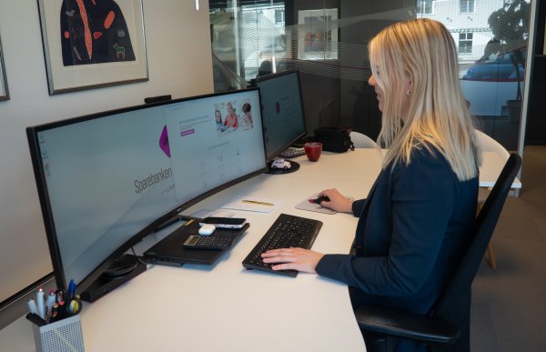 Alternativ tekst (for skjermleser): En kvinne sitter ved en kontorpult på et moderne kontor og jobber på en datamaskin med to store skjermer. På pulten ligger tastatur, mus, notatbok, kalkulator og kontorutstyr. Rommet har glassvegger og innrammede bilder på veggen.