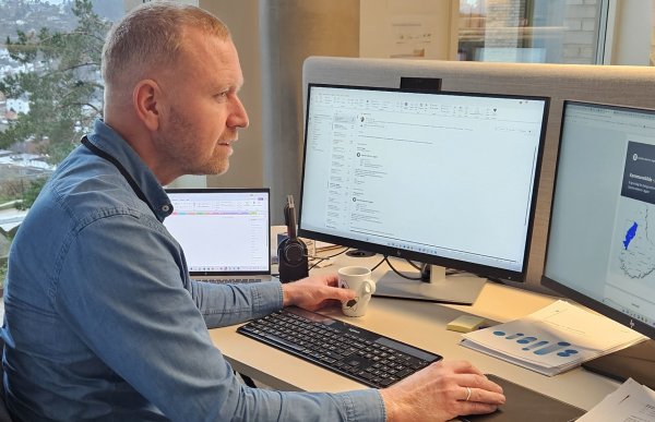 En mann sitter ved et kontorpult og jobber foran to store dataskjermer, med en bærbar PC ved siden av. Han holder en kaffekopp i den ene hånden og ser konsentrert mot skjermen.