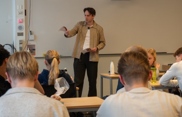 En mann står foran et klasserom med flere elever som sitter ved pulter vendt mot ham. Han holder et ark i den ene hånden og peker eller gestikulerer med den andre. Elevene følger med, noen med notatbøker og flasker på pulten.