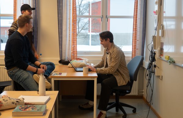En mann sitter ved et skrivebord foran en tavle og jobber på en bærbar PC. Han har en samtale med to unge gutter som står og sitter ved pulten ved siden av. Rommet er lyst, med store vinduer og skolebøker på bordet. Stemningen virker uformell. 