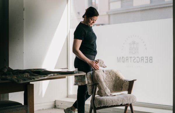 En møbeltapetserer står ved et vindu i verkstedet sitt og holder hånden på ryggen til en liten hund som sitter på en stol trukket i lys ull. Hun har sort T-skjorte på seg og mørkt hår i hestehale. Ved siden av står et bord med stoffprøver og materialer.