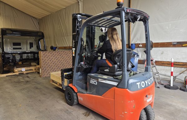 En svart og oransje truck hvor det står Toyato bakpå kjører fremover med en palle foran. Inni trucken sitter en dame med langt lyst hår.