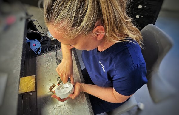 Kvinne med blond hestehale i blå arbeidsuniform filer på en hvit tannmodell ved en arbeidsbenk.