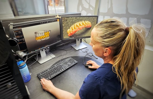 Kvinne med blond hestehale i blå arbeidsuniform sitter foran to dataskjermer og designer 3D-modeller av tenner.