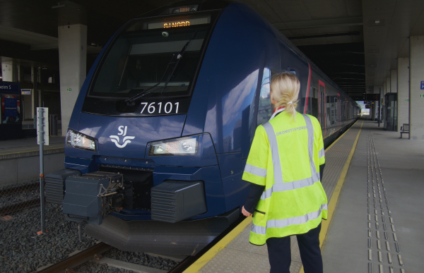 En kvinne i gul refleksvest står på perrongen med ryggen til kamera og kikker mot et tog. Toget har logoen SJ plassert foran.