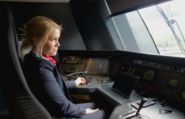 En kvinne i uniform sitter i en førerrstol i et tog. Foran seg har hun ulike spaker, skjermer og knapper. 