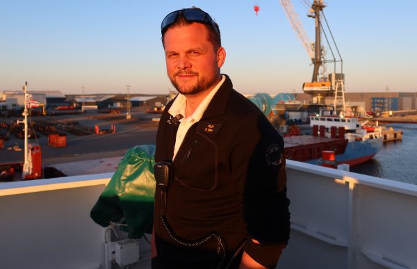 En mann i mørke klær. Han står på dekk på et stort skip med solnedgang i bakgrunnen. Bak han er et havneområde med kraner, containere og lasteskip i det fjerne.