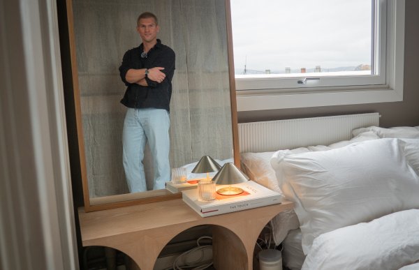 En mann står med hendene i kors, og vises i refleksjonen av et speil inne på et soverom. På sengen er det hvitt sengetøy, og ved siden av står det et nattbord i en bro-liknende form. 