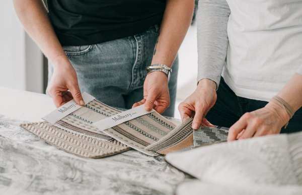 To personer holder og studerer ulike stoffprøver på et bord. Tekstilene har forskjellige mønstre og strukturer, og brukes til å velge trekk til møbler