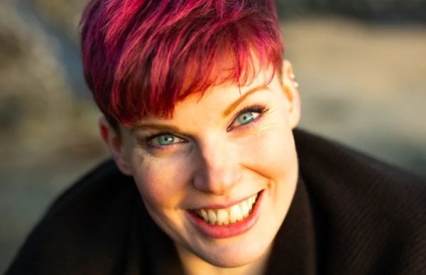 En kvinne med rødt, kort hår og blå øyne smiler inn i kamera.