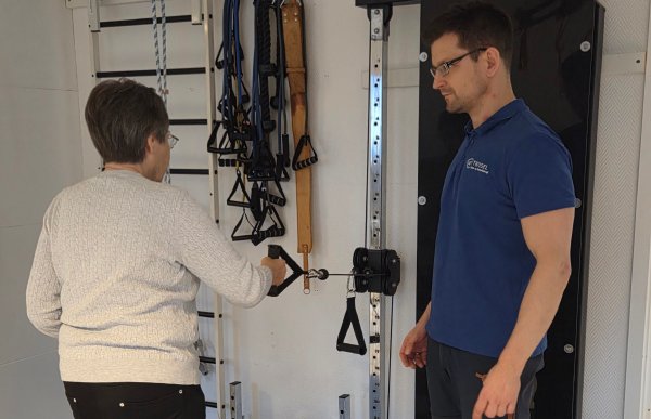En eldre kvinne står med ryggen til kamera og holder i et håndtak festet til et kabelapparat. Ved siden av henne står en mannlig behandler i blå poloskjorte og følger nøye med mens hun utfører øvelsen. Bak dem henger treningsstrikker og annet utstyr på veggen.