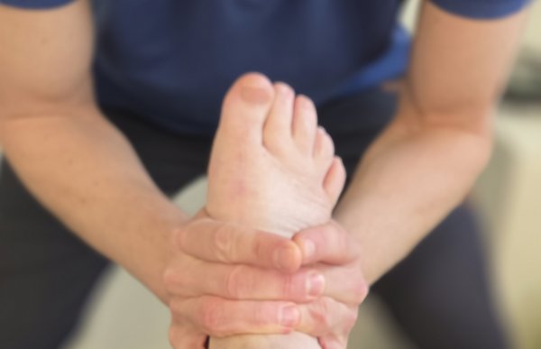 En behandler står bøyd over en pasients underben og holder rundt pasientens ankel med begge hender. Pasienten ligger på en benk med det ene beinet strukket ut mot behandleren. Fokus i bildet er på foten og hendene som undersøker eller mobiliserer ankelen.