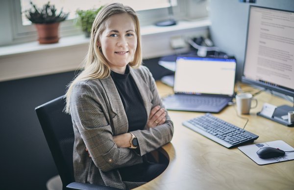 En kvinne med langt blondt hår sitter ved et skrivebord på kontoret sitt. Hun har armene i kors, smiler og ser inn i kamera. På pulten står to dataskjermer, tastatur, kaffekopp og kontorutstyr.