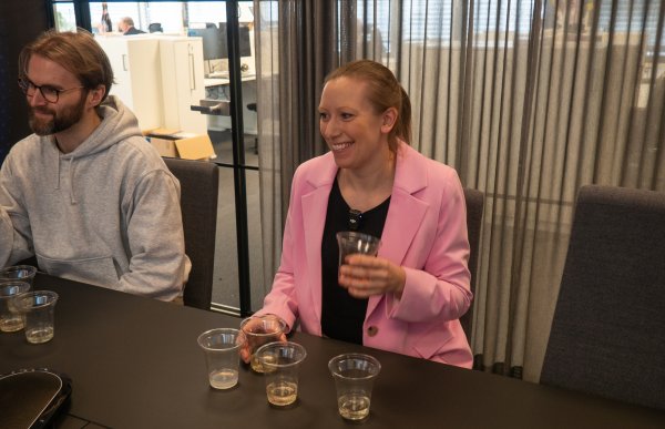 En kvinne sitter ved et møtebord sammen med en annen person på et kontor. Hun har på seg en rosa blazer og holder et lite glass i hånden. På bordet står flere små glass med innhold, og situasjonen fremstår som en smaking eller gjennomgang i jobbsammenheng. Kontorlokalet har glassvegger og gardiner i bakgrunnen.