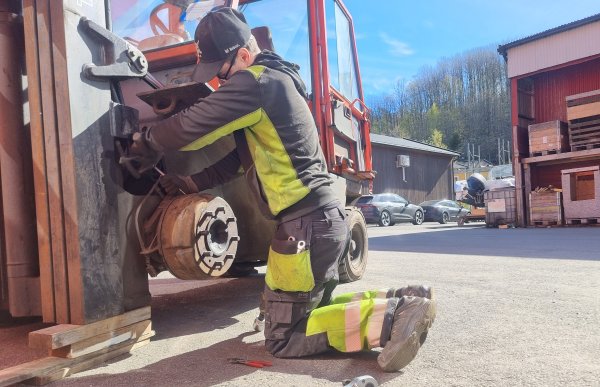 En mann er foran en rød gaffeltruck og arbeider på masten. Han bruker arbeidsklær med synlige refleksfelt. Han har verktøy liggende rundt seg på bakken. 