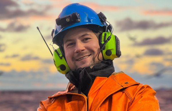 En mann i oransje arbeidsklær, blå hjelm og grønne hørselvern. Han er ute.