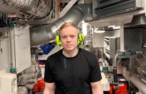 En mann står i et trangt maskinrom omgitt av store motorer, rør, ventilasjonssystemer og teknisk utstyr. Han har på seg hørselsvern og arbeidsklær, og ser rett mot kameraet. Rommet er tettpakket med maskineri og metallrør i ulike størrelser.
