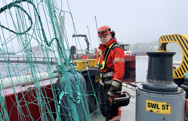 En mann i røde arbeidsklær, hjelm og redningsvest står på dekk på en arbeidsbåt ute på sjøen. Rundt ham henger grønne tau og notliner, og i bakgrunnen sees oppdrettsanlegg, arbeidsutstyr og gråvær. Han holder en verktøykasse i hånden og står mellom store vinsjer og dekksutstyr.