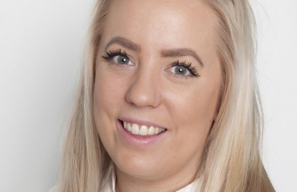 Et nært portrettbilde av en ung kvinne som smiler i kameraet. Hun har langt, lyst hår. Hun har på seg en hvit genser med en hvit skjortekrage. 