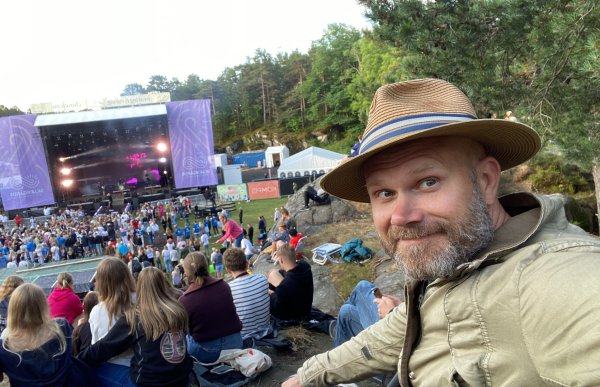 En mann med hatt og skjegg i lys jakke står ute foran en stor scene og mange ungdommer som er publikum