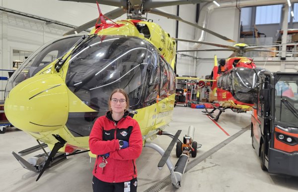 En kvinne med briller og rød arbeidsjakke står med armene i kors foran et stort, gult helikopter i en lys hangar. Et mindre rødt og gult helikopter og et svart og rødt kjøretøy står lenger bak.