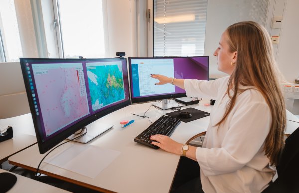 En kvinne sitter på et lyst kontor foran to store dataskjermer. Det er flere vinduer med forskjellige kartutsnitt, farger og værdata, og hun peker på skjermen.