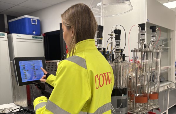 En kvinne i gul arbeidsjakke står i en kontrollrom og ser på en dataskjerm