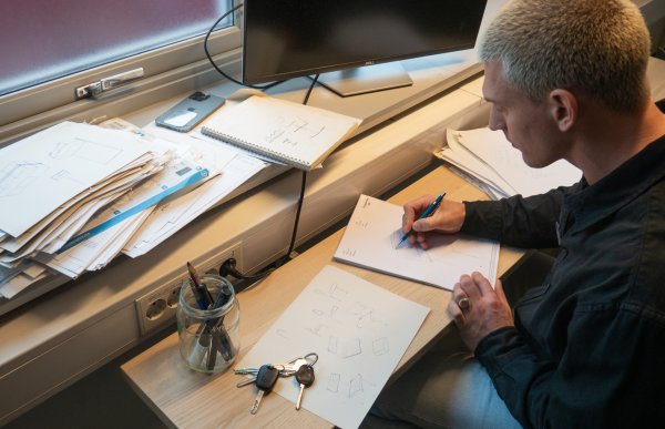 En mann sitter med ryggen mot kameraet og tegner på noe papir på pulten foran seg. Han skisserer noen stoler, og det ligger skisser over hele pulten. Det står også en avslått PC skjerm på pulten.