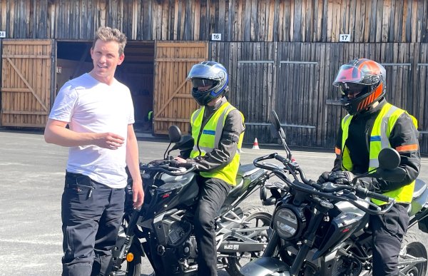 En trafikklærer i hvit t-skjorte står ved siden av to elever som sitter på hver sin motorsykkel. Elevene har på seg hjelm og gule refleksvester. Bildet er tatt på en øvingsplass foran en trebygning med nummererte porter.