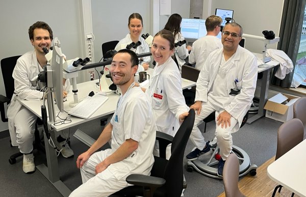 En gruppe på seks ansatte i hvite frakker sitter ved mikroskoper og PC-er i et lyst laboratoriekontor. De smiler mot kameraet i et aktivt, faglig arbeidsmiljø.