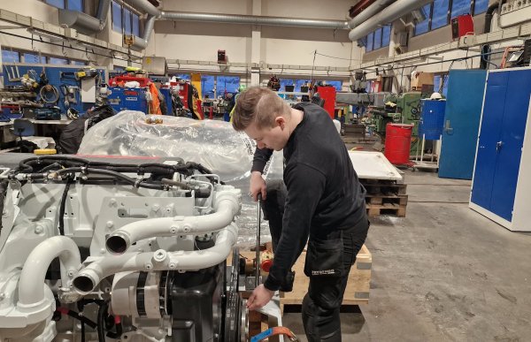 Mann med brunt hår og svart genser står foran en motor og skrur på denne