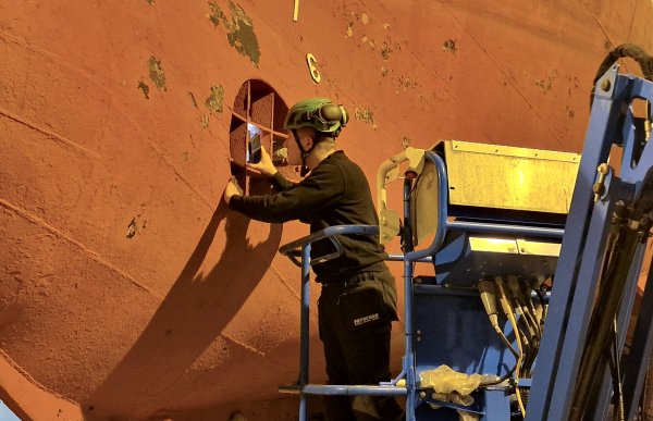 Mann med svart bukse og genser står i en stige og bruker mobilen til å lyse inn i et rom på en båtside. Han har hørselvern og hjelm på seg.