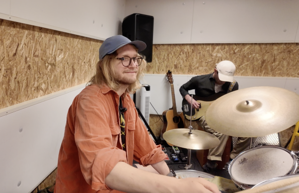 En person med caps, skulderlangt hår og briller sitter bak et trommesett i et musikkrom. Bak trommesettet sitter en annen person med caps på, og spiller på en bass. 