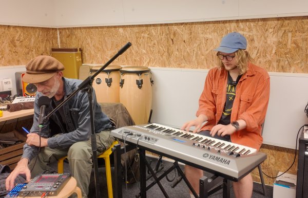 To personer sitter i et musikkrom. Den ene spiller på et Yamaha-keyboard mens den andre sitter foran en mikrofon og justerer en lydmodul. Bak dem står trommer, forsterkere og forskjellig musikkutstyr stablet langs veggen.