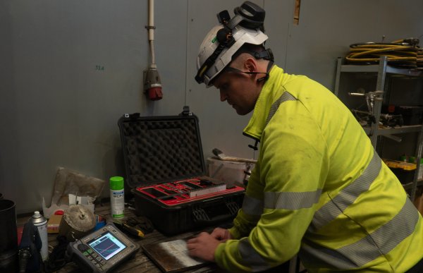 En mann i gul refleksuniform, hjelm og hørselsvern holder en liten scanner mot en stålbit på bordet foran seg. Det ligger en åpen utstyrskoffert foran han. Han kikker på en skjerm som viser tre ulike grafer.  