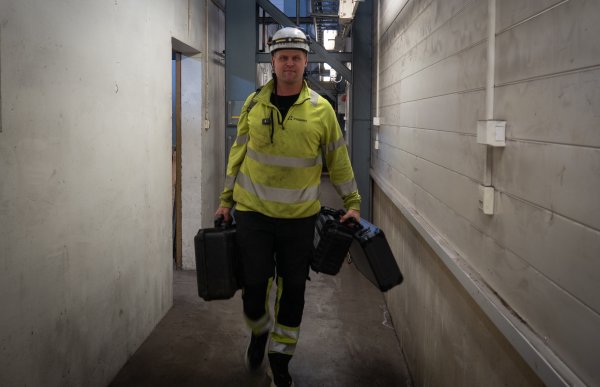 En mann i gult arbeidstøy og byggehjelm på hodet kommer gående gjennom en industriell korridor. Han har tre store svarte utstyrskofferter i hendene.  