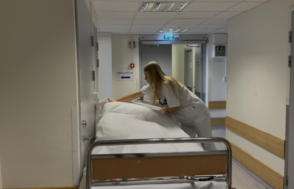 En kvinne med blondt, langt hår iført hvite sykehusklær rer opp en sykehusseng i en korridor. Hun lener seg over sengen mens hun ordner sengetøyet. Rundt henne er det lyse sykehusvegger, dører og skilt i gangen.