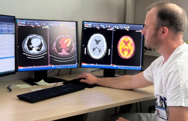 Mann i hvit t-skjorte sitter ved en pult. Det står tre pc-skjermer foran ham. Pc-skjermene viser bilder av kroppen.