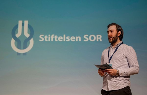 En mann står på en scene og holder et ark i hendene. Han har skjorte, mørk bukse og bærer mikrofon på hodet. Bak ham vises en stor skjerm med teksten «Stiftelsen SOR» og en logo.