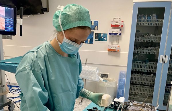 En person ikledd operasjonsklær står ved et bord med instrumenter i en operasjonsstue. Hun har på munnbind og grønt hårnett.