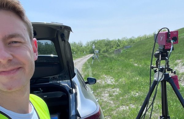 En mann i gul refleksvest tar en selfie ute på et jorde, med bilbakluken åpen bak seg. Foran bilen står et stativ med et rødt hyperspektralt kamera som peker ut over det åpne landskapet.
