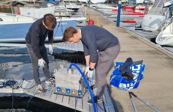 To personer står på en brygge i en småbåthavn og jobber med et stort måleinstrument koblet til blå kabler som går ned i vannet. Rundt dem ligger flere seilbåter fortøyd, og sola skinner over havna.