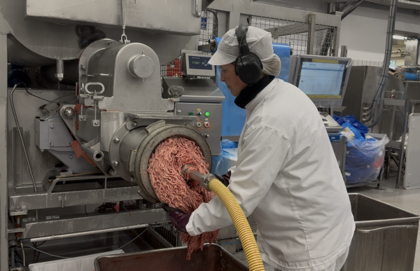 En kvinne i hvit arbeidsfrakk, hårnett og hørselsvern står ved en stor kjøttkvern i et produksjonslokale. Hun håndterer kjøtt som kommer ut av maskinen. Rundt henne er det industrielle maskiner og skjermer.