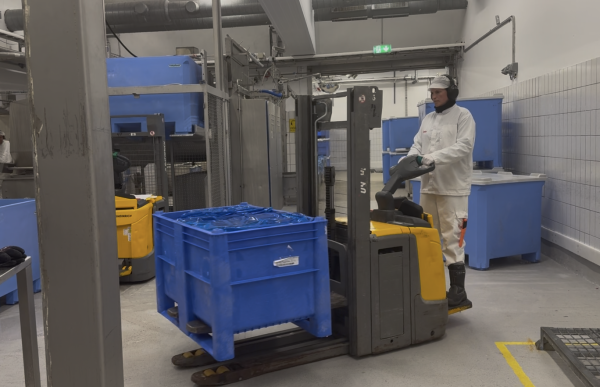 En kvinne i hvite arbeidsklær, gummistøvler og hørselsvern kjører en truck med en stor blå kasse i et produksjonslokale. Rundt henne er det maskiner, kasser og annet utstyr i et industrielt miljø.