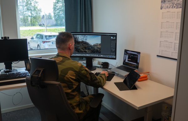 En person i militæruniform sitter ved et skrivebord og redigerer bilder på en stor skjerm. På pulten står flere skjermer og en laptop, og et vindu ved siden av viser en parkert bil utenfor.