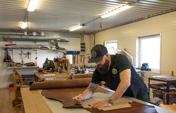 Mann med svart t-skjorte og caps står over et arbeidsbord. Det ligger et skinn der og han jobber med å ta mål.