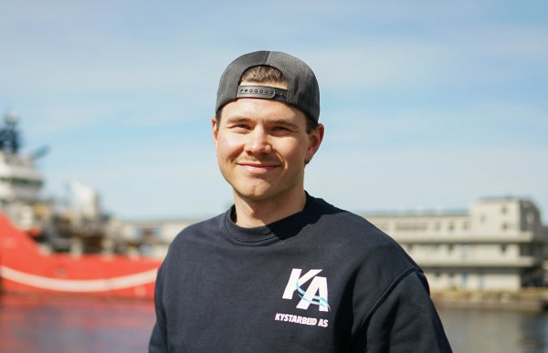Ung mann med skyggelue bak fram, smiler til kameraet. I bakgrunnen er det en stor båt og sjø.