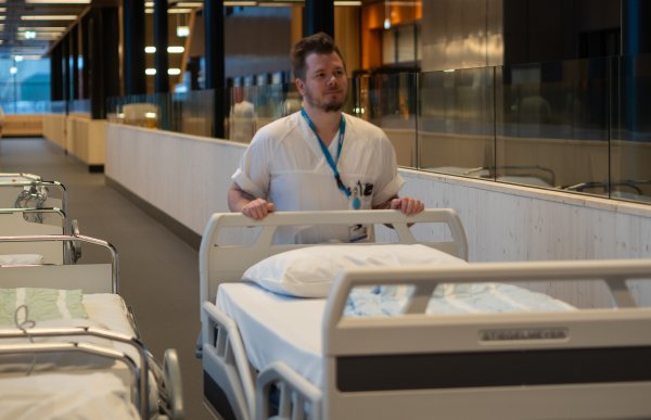 En mann går i en bred korridor inne i et sykehus og skyver en tom sykehusseng foran seg. Han har på seg hvite arbeidsklær og et adgangskort rundt halsen. På begge sider av korridoren står flere senger, og området er godt belyst.