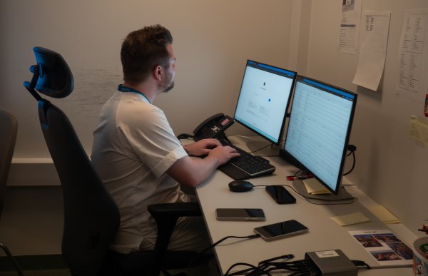 En mann sitter ved et skrivebord på et kontor og jobber på en datamaskin med to skjermer. Han skriver på tastaturet og ser på skjermen foran seg. På pulten ligger mobiltelefoner, notatlapper, kabler og kontorutstyr. Rommet er lite og enkelt innredet, med papirer festet på veggen.