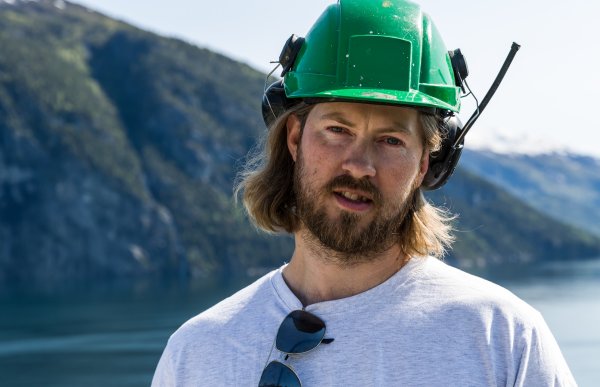 Mann med brunt hår og hvit t-skjorte og grønn anleggshjelm med hørselsvern. Fjord og fjell i bakgrunnen.